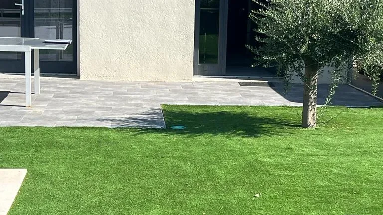 Façade enduite beige donnant sur une terrasse en dalles grises, une pelouse et un olivier